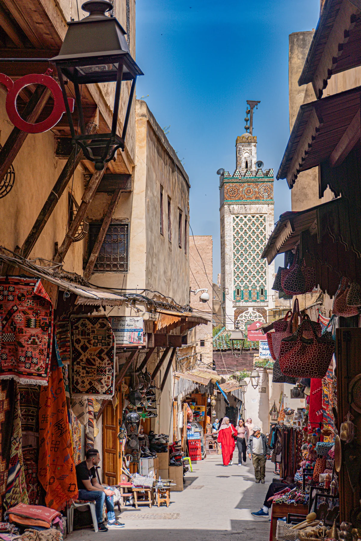 Smalle marktstraat in een Marokkaanse medina met tapijten en tassen te koop, uitzicht op een versierde minaret