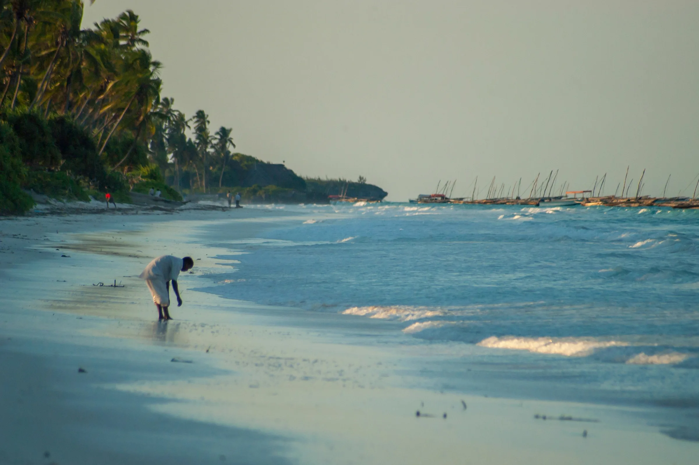 Persoon die bij avondlicht langs de vloedlijn loopt, met traditionele zeilboten voor de kust van Zanzibar